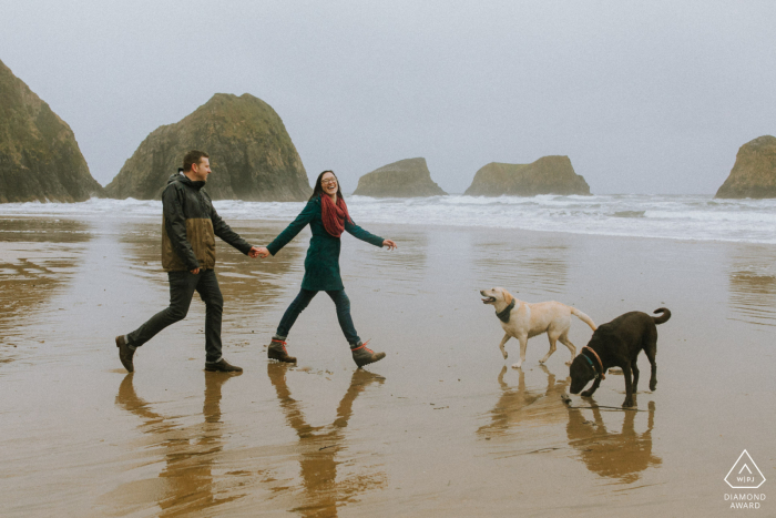 Una pareja comprometida paseando a sus perros en una playa lluviosa del Parque Estatal Ecola para una sesión de retrato