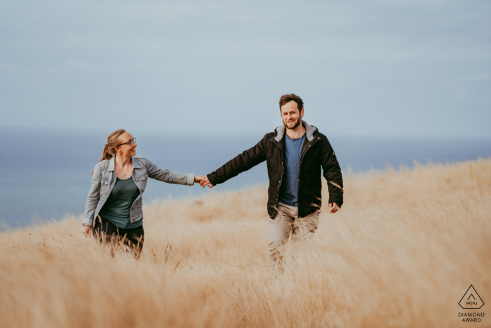 Un Christchurch NZ Couple marchant main dans la main dans les hautes herbes au bord de la plage au cours de leur séance photo avant le mariage