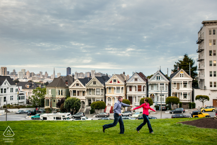 N. CA sessão de fotos de noivado em São Francisco de um casal correndo no gramado em frente ao Painted Ladies