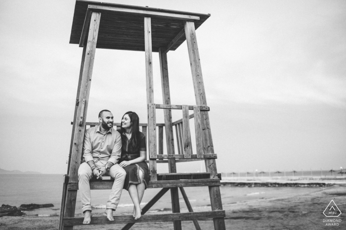 Atenas, Grecia pareja sentada en la playa torre de salvavidas riendo en esta foto de compromiso en blanco y negro