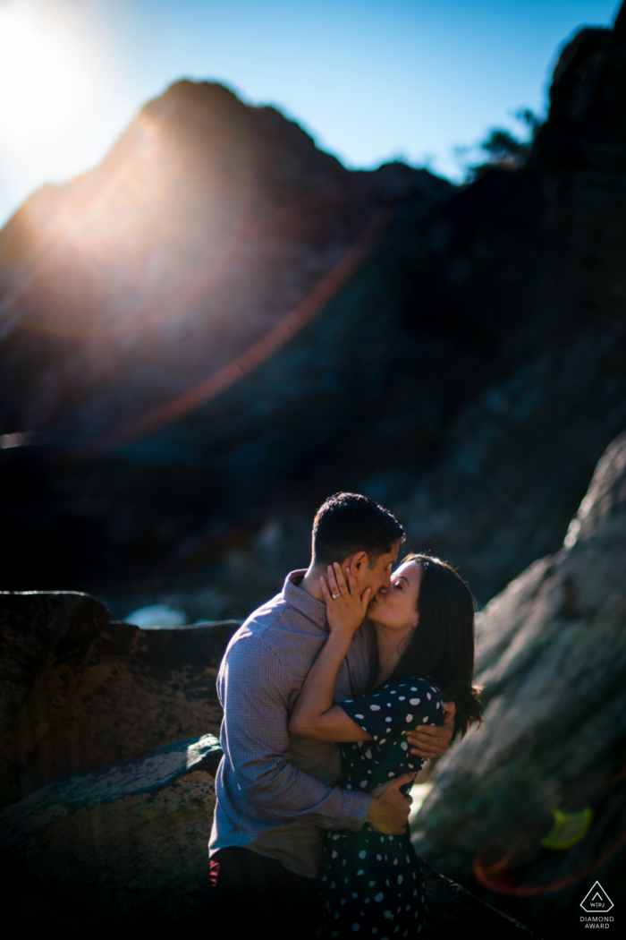 Virginia engagement portrait with a posed couple at Great Falls VA with a kiss in front of a large rock, lens flare accents the upper left corner