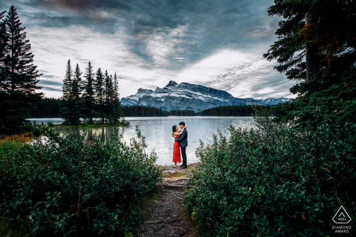 Two Jacks Lake, AB, Canadá, mini sesión de fotografía de pareja en la playa antes del día de la boda mientras se abrazan al aire libre en la naturaleza