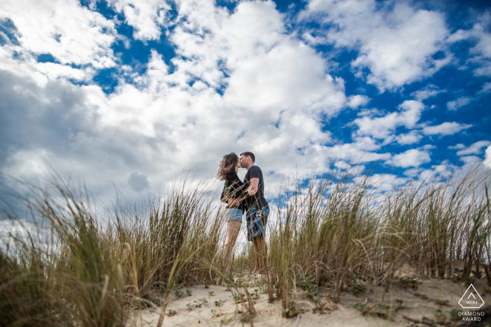 Bassin d Arcachon, France mini beach couple photography session before the wedding day with A kiss on a sand dune under blue skies with clouds Bassin d Arcachon, France mini beach couple photography session before the wedding day with A kiss on a sand dune under blue skies with clouds