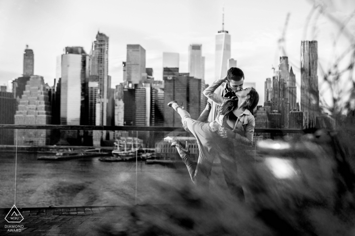 Mini-sessão de fotos de casal no Brooklyn Bridge Park, em Nova York antes do dia do casamento em um abraço de dançarinos com o horizonte da cidade atrás deles