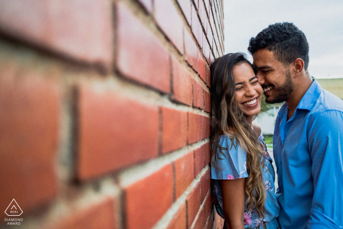 Sessão fotográfica pré-casamento do casal em Maceió sorrindo em pé contra uma parede de tijolos