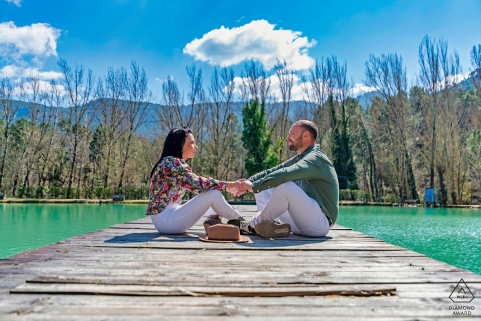 Jaen Paar Verlobungsfotosession im Hotel Noguera de la Sierpe in Arroyo Frío auf dem Holzdock am Wasser, sitzend und Händchen haltend