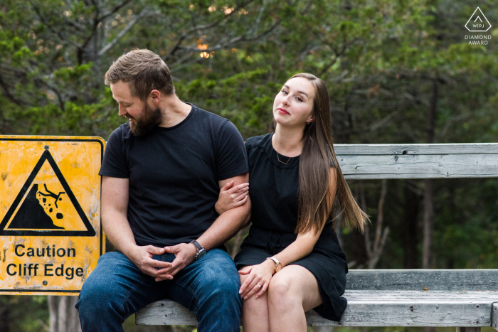 Westport Lookout Point, Ontario Fine Art Engagement Image for a couple sitting on a bench next to a sign reading Caution cliff edge 