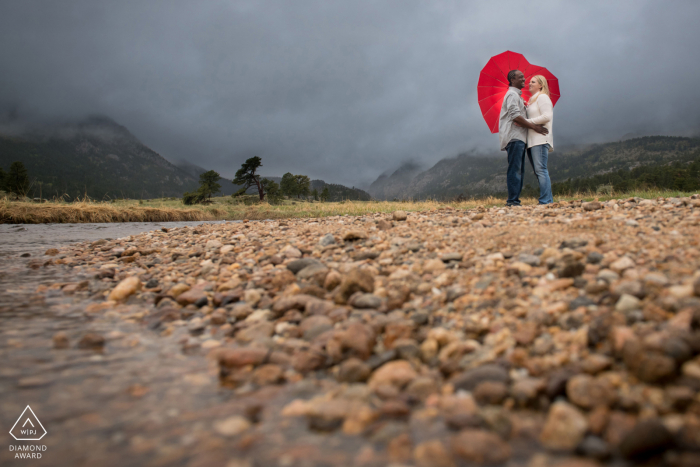 Rocky Mountain National Park Fine Art Engagement Image for a Couple under heart umbrella on a rainy day by a river in a large valley 