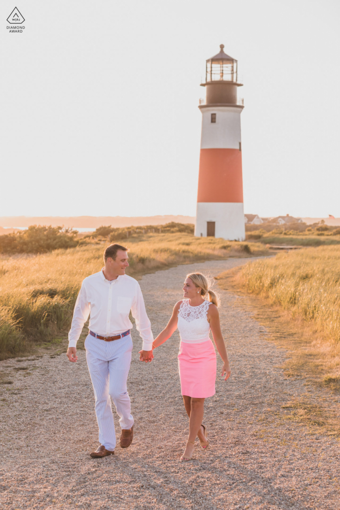 Sankaty Lighthouse e-session sull'impegno ambientale sull'isola di Nantucket, MA di una coppia che fa una passeggiata con un faro dietro di loro