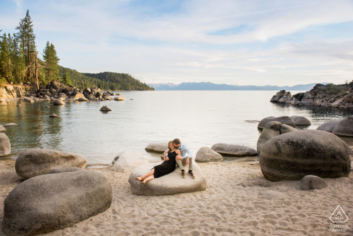 Il vero amore fidanzamento ha posato ritratto a Lake Tahoe, NV che cattura un paio di abbracci seduti su una roccia con la distesa dell'acqua sullo sfondo