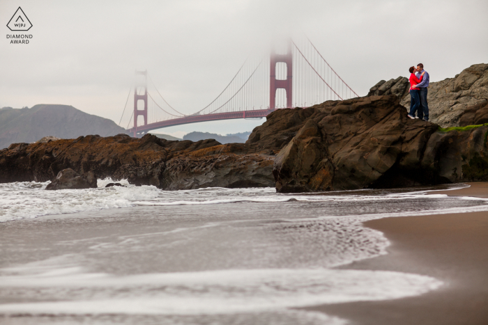 Prawdziwa miłość pozowany portret zaręczynowy w San Francisco ukazujący kochanków na plaży, z zasłoną mgły zakrywającą most Golden Gate w tle