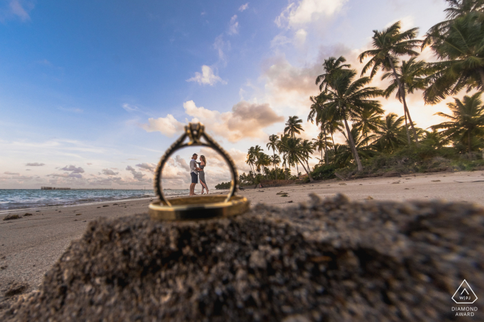 Sessão de retratos pré-casamento True Love em Maragogi ilustrando um casal em frente ao mar e fotografado através de anéis na areia