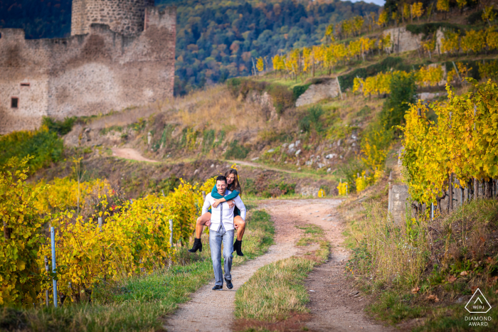 Photo de fiançailles du château de Kaysersberg, en Alsace, montrant la facilité naturelle de leur relation alors que le monsieur porte sa fiancée sur un chemin de campagne loin du château