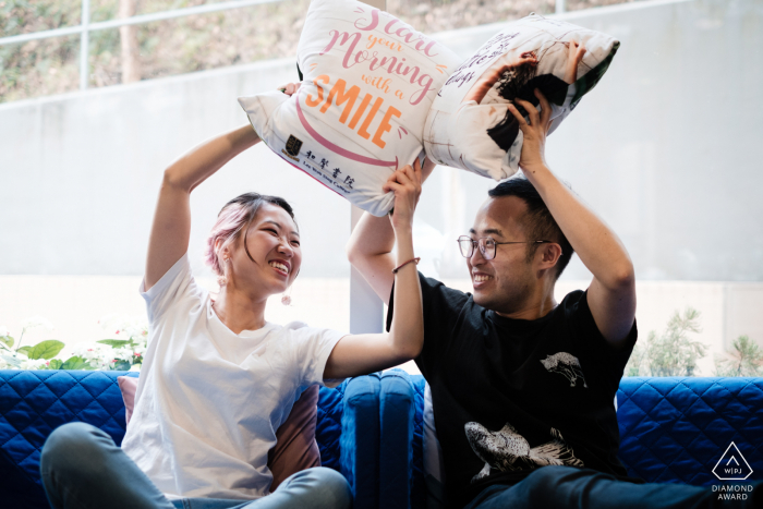 Retratos pre-boda de la Universidad China de Hong Kong para una divertida pareja que tiene una pelea de cojines en un día que vuelven a visitar el campus universitario donde se conocieron y el nombre y el emblema de la Universidad se pueden ver en uno de los cojines.