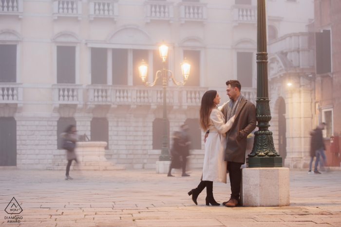 Ein italienischer Hochzeits-, Elopement- und Verlobungsfotograf hat dieses künstlerische, urbane Porträt dieses Paares aus Venedig erstellt, das im Nebel an einem Laternenpfahl lehnt