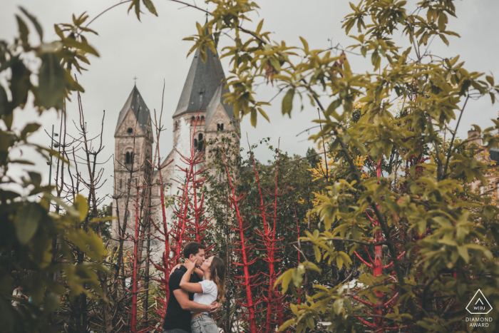 A Metz couple poses during a France lifestyle portrait photography session under the tall branches with a tall building in the distance A Metz couple poses during a France lifestyle portrait photography session under the tall branches with a tall building in the distance