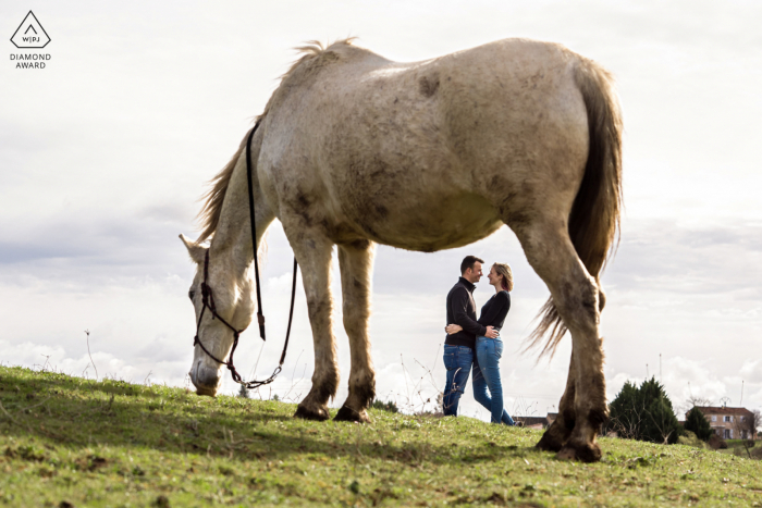 Albi post proposal photography showing a smiling France couple standing far behind their horse grazing on field grass