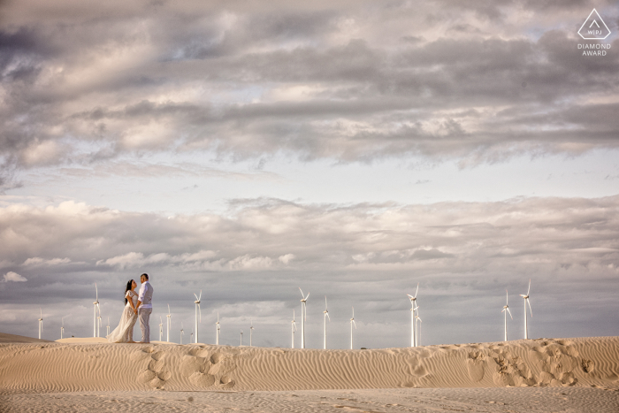 Dunas de Salinas fidanzamento. Una giovane coppia di Rio Grande do Sul in posa durante una pittoresca sessione di ritratti prima del matrimonio in spiaggia con mulini a vento sullo sfondo