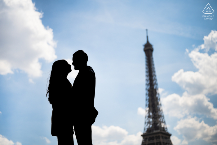 Engagement Photos Paris. A French couple pose against the blue sky, clouds and a silhouetted Eiffel Tower 