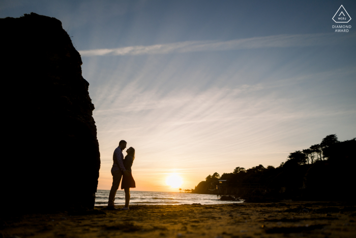 Ein Fotoshooting vor der Hochzeit in Pornic, Loire Atlantique, Frankreich, das zu einer Silhouettenaufnahme eines Paares führte, das sich bei Sonnenuntergang zwischen Felsen am Strand küsst