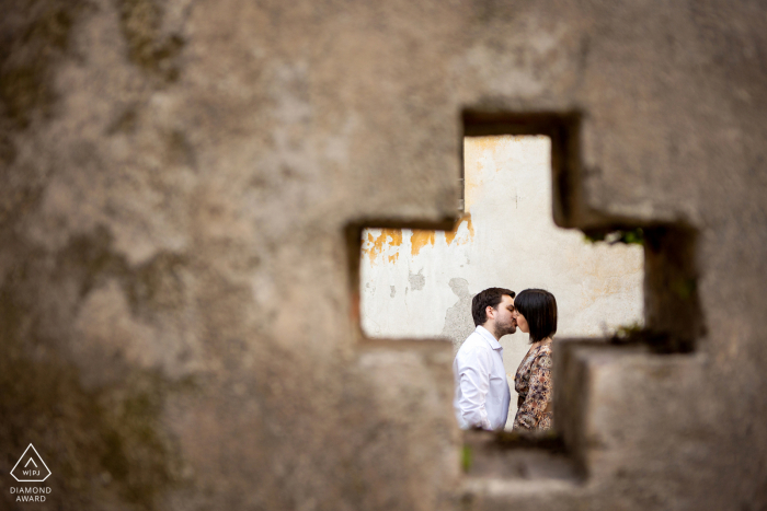 An after the marriage proposal Italy engagement photograph at Brescia Castle with kissing lovers shot through a cross shaped window opening