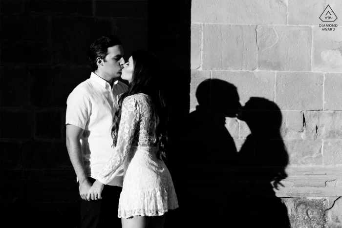 Ein Verlobungsfoto nach dem Heiratsantrag in Italien in der Kathedrale von Arezzo für ein Paar, das in einem Kuss zwischen Licht und Schatten in der Nähe der Kathedralenmauer gefangen ist