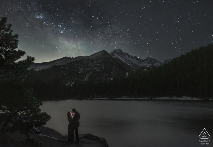 A Colorado modern couple poses for outdoor, night announcement portrait of their RMNP engagement at Bear Lake under the rising Milky Way lit with flash, and a single exposure