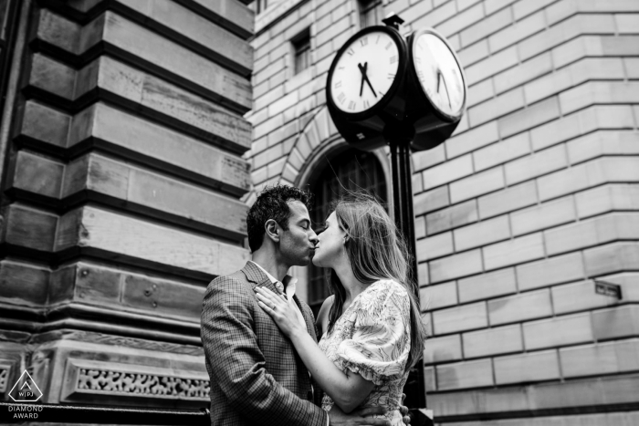 An Old Montreal outdoor portrait shows a unique way this newly engaged couple is announcing their Canada engagement as they are kissing below an antique street clock surrounded by old stone buildings 