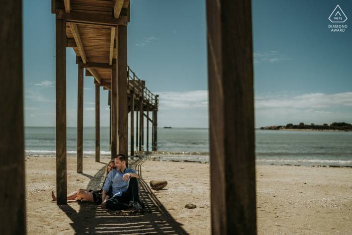 Un couple moderne de l'île d'Aix pose à l'ombre sous la jetée de la plage pour un portrait d'annonce de leurs fiançailles en Charente-Maritime