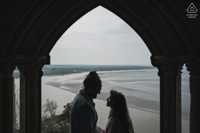 Un couple de Normandie nouvellement fiancé pose dans une arche pour un portrait d'annonce de fiançailles du Mont Saint Michel sur une île de marée et une commune de Normandie, France