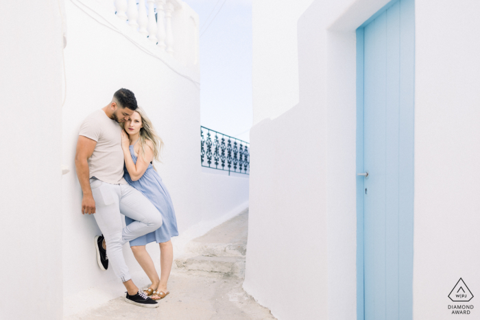 Santorini couple posing for a creative engagement announcement shot in Greece as they are hugging and leaning against greek white walls 