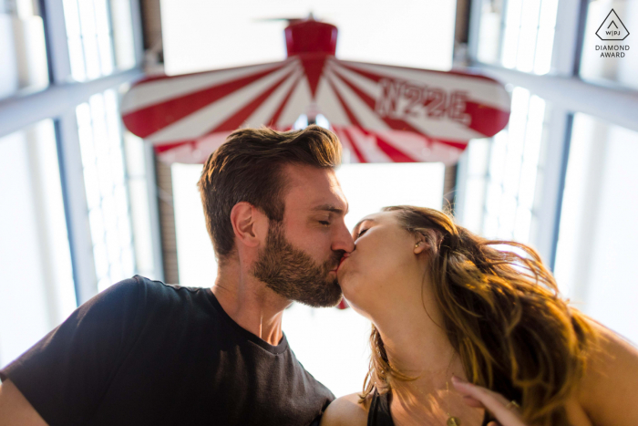 Washington DC Lifestyle-Engagement-Fotografie im Air and Space Museum für ein Paar, das sich mit dem Flugzeug über dem Kopf küsst