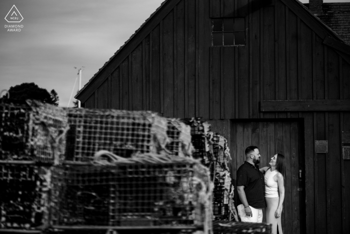 Rockport, Massachusetts, nicht traditionelles Lifestyle-Engagement-Porträt eines Paares, das an den Fischerdocks in der Nähe eines Stapels Hummertöpfe posiert