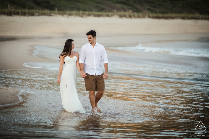 Fotografia de noivado de estilo de vida de Niterói de um casal andando na areia e água ao longo da costa do oceano