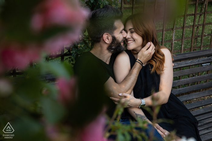 Fotografia de noivado de estilo de vida de Nancy de um casal francês abraçado em um banco de parque perto de algumas flores cor de rosa