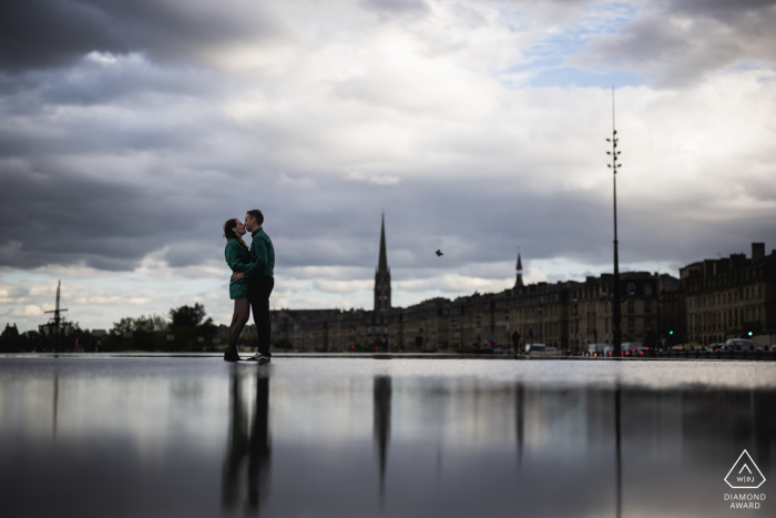 A France wedding photographer created a reflection engagement portrait for a Bordeaux couple kissing under a cloudy, stormy sky 