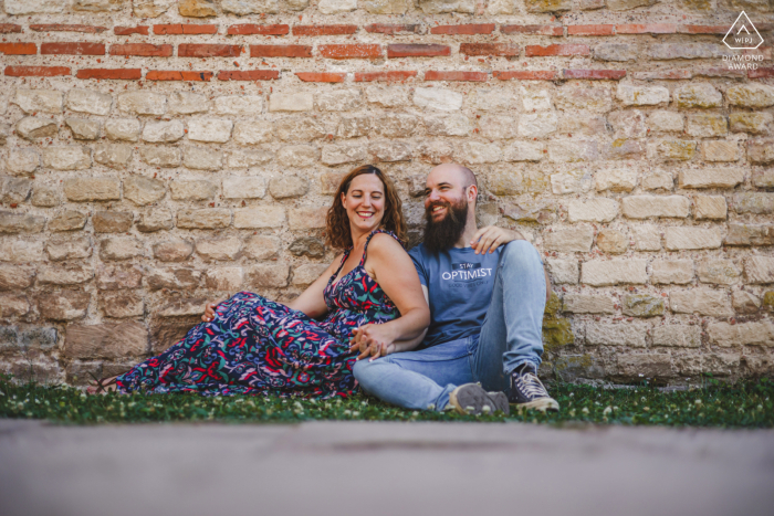 Frente a una pared en Metz, Francia, la pareja sonriente está sentada en el césped frente a una pared de ladrillos.