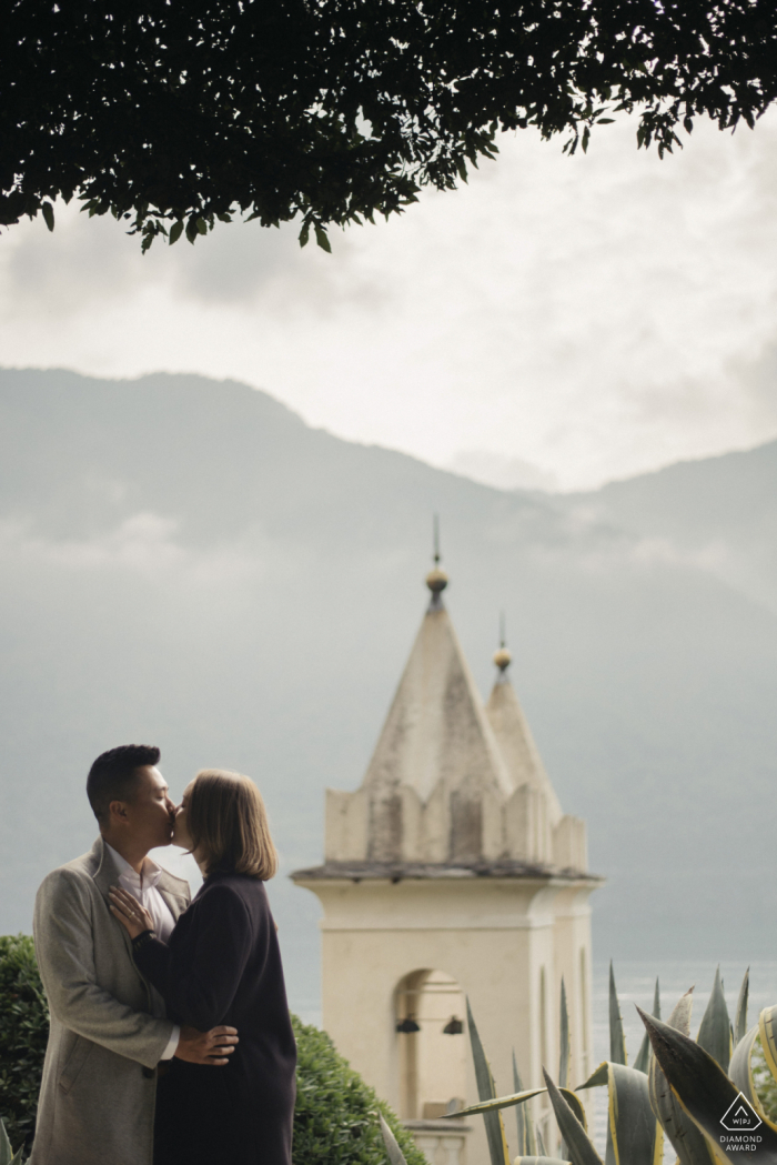 Das Paar teilt eine romantische Umarmung, ihre Lippen treffen sich, während der Sonnenuntergang eine sanfte Farbpalette über den Blick auf den Comer See von der Villa Balbianello wirft