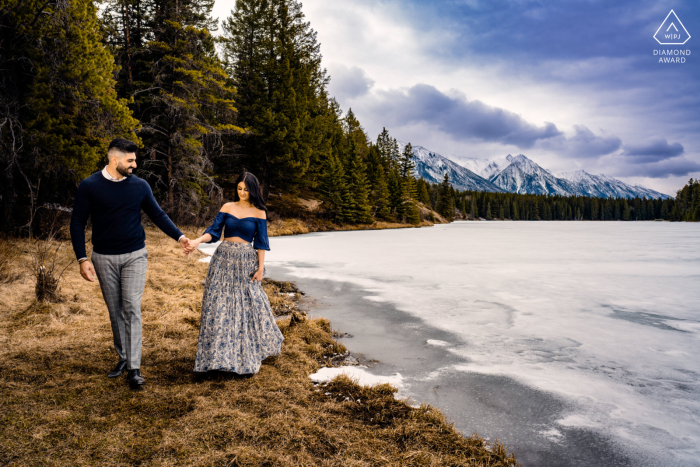 An engaged couple is featured in a portrait against a shoreline backdrop, embracing while walking hand-in-hand The picturesque scenery of Banff, Canada provides the perfect backdrop for a glamorous couple's photo session as they walk the shore holding hands