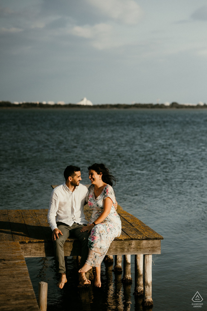 A young couple pause together for an engagement portrait on a wooden dock overlooking a pond in Mauguio