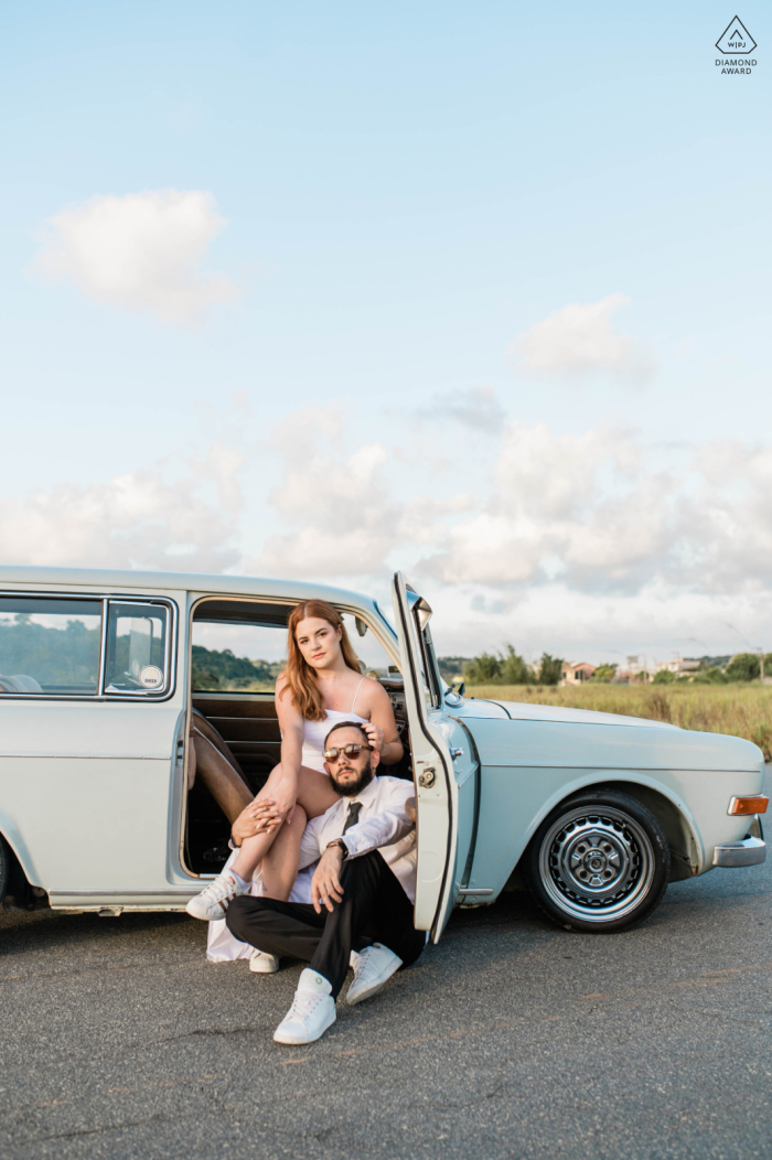 A essência da velha Hollywood com um carro antigo, enquanto um homem se senta no chão e uma mulher posa dentro do carro durante uma sessão de fotos de noivado