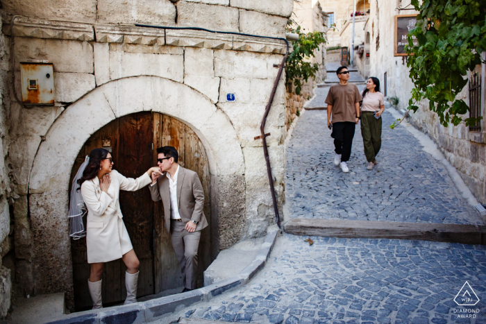 In a pre-wedding photo session in Cappadocia, Turkey, at Goreme, a couple is pictured on a cobblestone road, where the groom affectionately kisses the bride's hand as another couple walks towards them