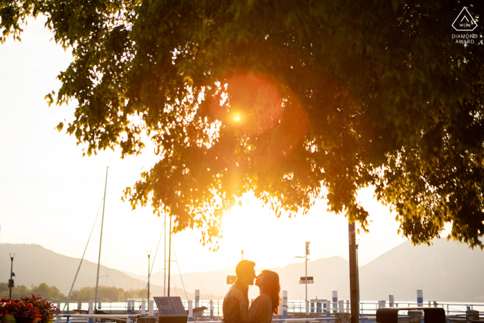 A couple is captured in silhouette at Iseo Lake in Italy, standing near boats by the water, with sunlight filtering through the trees above them
