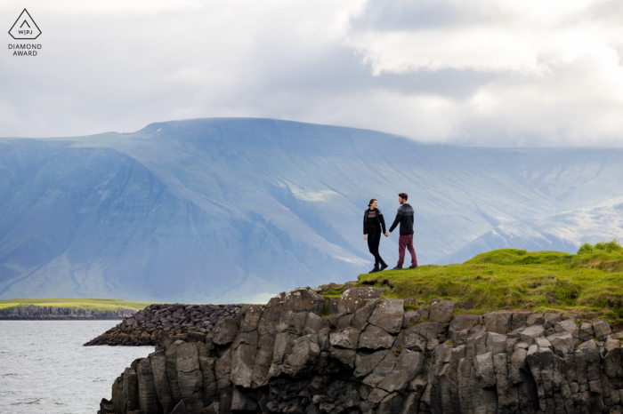 Fine art couple portrait by a Reykjavik wedding photographer, capturing a couple walking and holding hands in front of a stunning landscape of water and mountains in Iceland.