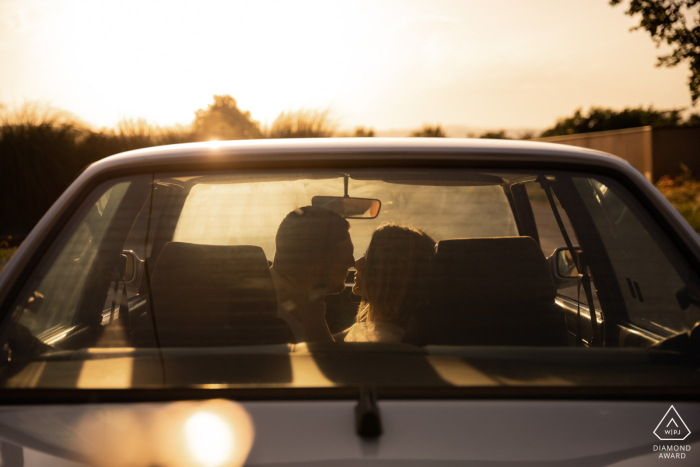 During their engagement portrait session at Valence, France, the loving couple sits in an old car at sunset, exchanging kisses with the romantic sky as their backdrop.