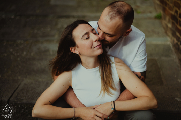An Engagement Portrait in the Charming Streets of Siena A couple poses for an engagement portrait during a relaxed session in the romantic setting of Siena, Tuscany. The man embraces his fiancée from behind in a sweet kiss.