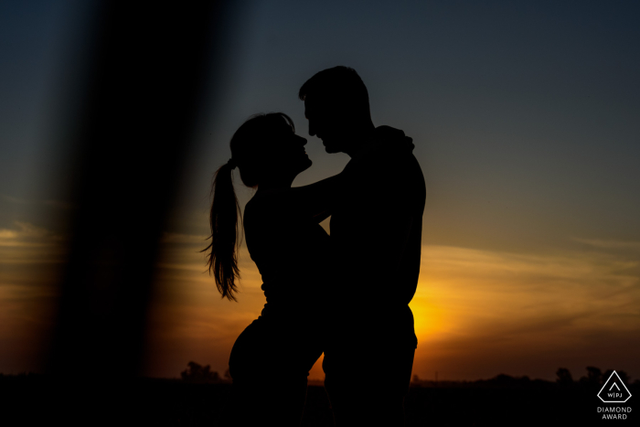 A skillful capture by a WPJA photographer in Zavalla, Santa Fe, Argentina. A couple, silhouetted in a vibrant sunset sky, embrace in a cornfield during their pre-wedding portrait session. 