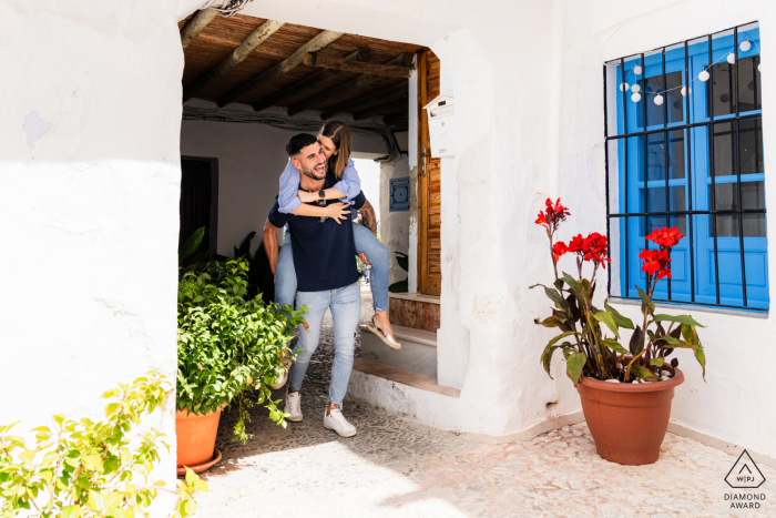 In this image from a pre-wedding portrait session by a WPJA professional in Frigiliana, Málaga, Spain, the couple playfully engages as she rides piggyback on him in the charming white painted village. 