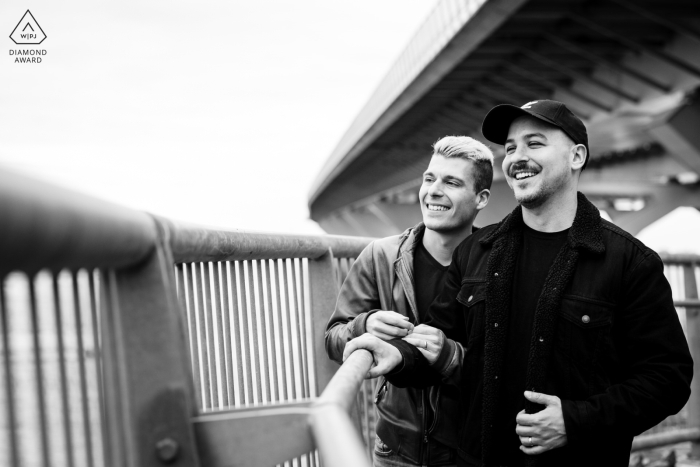 A professional WPJA photographer captures a black and white image of an engaged couple in Montreal, Quebec as they stand on a bridge, gazing out over the railing.