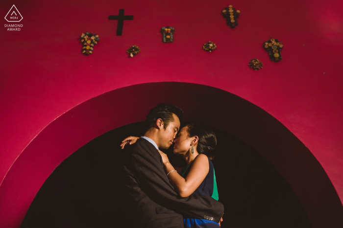 Engaged couple captured in a romantic kiss, framed by iconic red wall and crucifixes in San Miguel de Allende pre-wedding portrait session in Guanajuato.